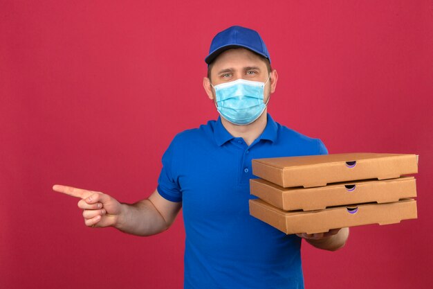 Repartidor joven vistiendo polo azul y gorra en máscara médica sosteniendo una pila de cajas de pizza apuntando con el dedo hacia el lado de pie sobre fondo rosa aislado
