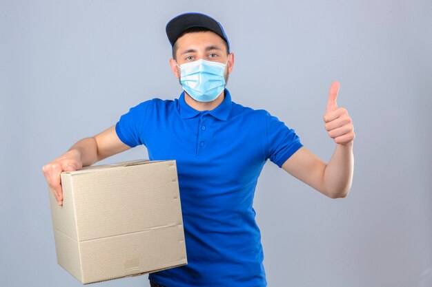 Repartidor joven vistiendo polo azul y gorra en máscara protectora médica de pie con caja de cartón mostrando pulgar mirada segura sobre fondo blanco aislado