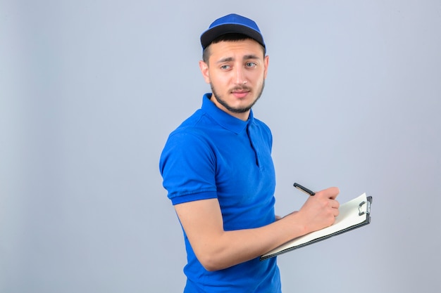 Repartidor joven vistiendo polo azul y gorra mirando aburrido de pie con el portapapeles y escribiendo sobre fondo blanco aislado