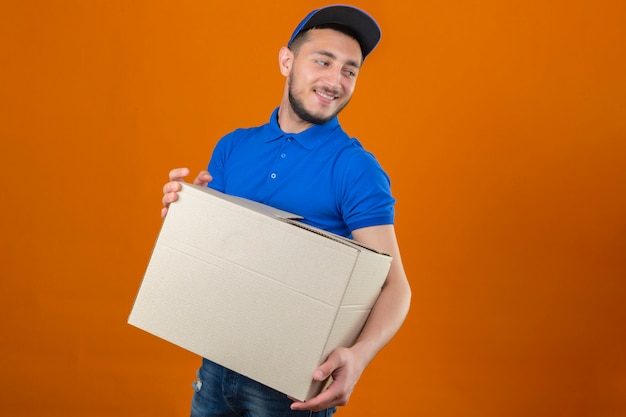 Repartidor joven vistiendo polo azul y gorra de pie con caja de cartón mirando hacia el lado con una sonrisa en la cara sobre fondo naranja aislado