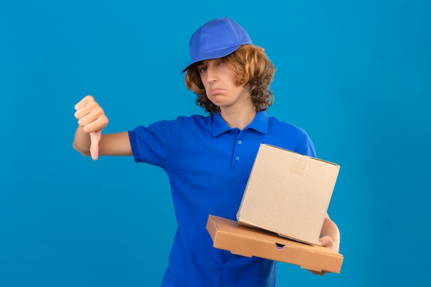 Repartidor joven vistiendo polo azul y gorra sosteniendo cajas de cartón mostrando el pulgar hacia abajo mostrando desagrado de pie sobre fondo azul aislado