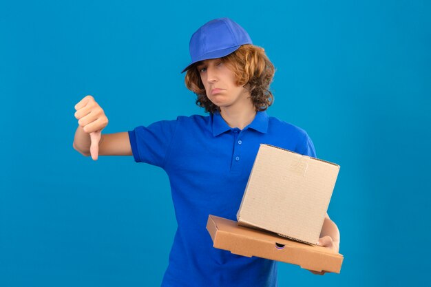 Repartidor joven vistiendo polo azul y gorra sosteniendo cajas de cartón mostrando el pulgar hacia abajo mostrando desagrado de pie sobre fondo azul aislado