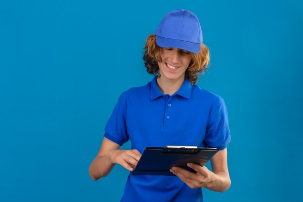 Repartidor joven vistiendo polo azul y gorra sosteniendo portapapeles mirándolo con una sonrisa en la cara sobre fondo azul aislado