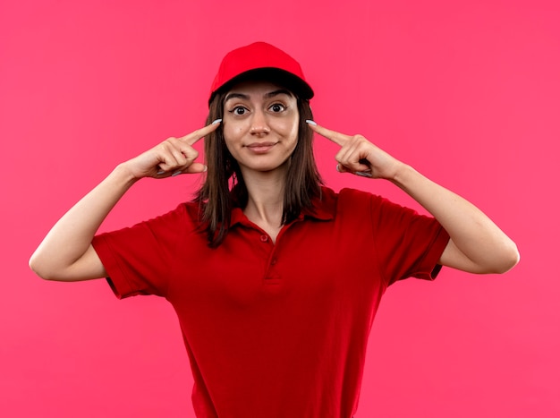 Repartidor joven vistiendo polo rojo y gorra apuntando a sus sienes mirando confidetn sonriendo concentrándose en una tarea de pie sobre la pared rosa