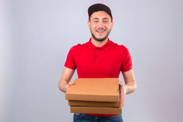 Repartidor joven vistiendo polo rojo y gorra estirando una pila de cajas de pizza sonriendo amable sobre fondo blanco aislado