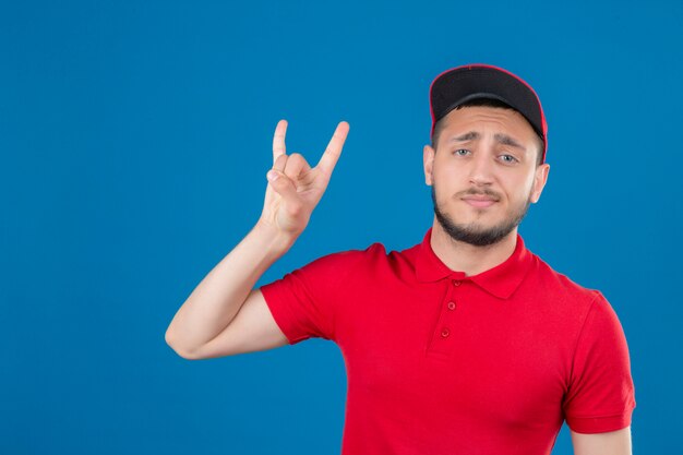 Repartidor joven vistiendo polo rojo y gorra haciendo símbolo de rock mirando confiado sobre fondo azul aislado