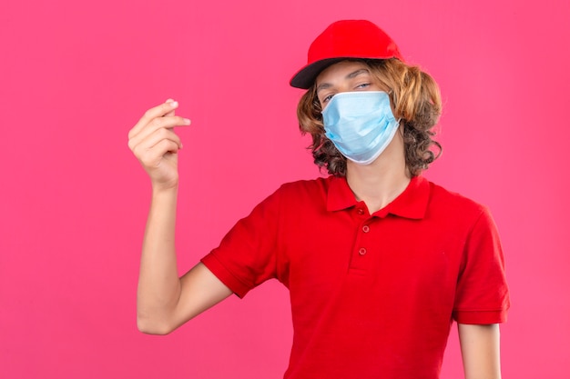 Repartidor joven vistiendo polo rojo y gorra en máscara médica haciendo un gesto de dinero mirando confiado sobre fondo rosa aislado