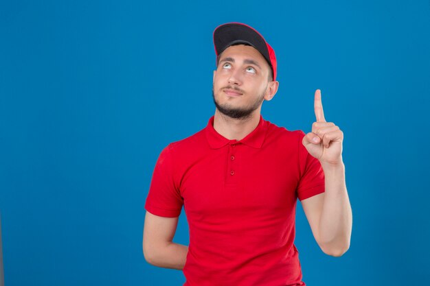 Repartidor joven vistiendo polo rojo y gorra mirando confiado con el dedo apuntando hacia arriba sobre fondo azul aislado
