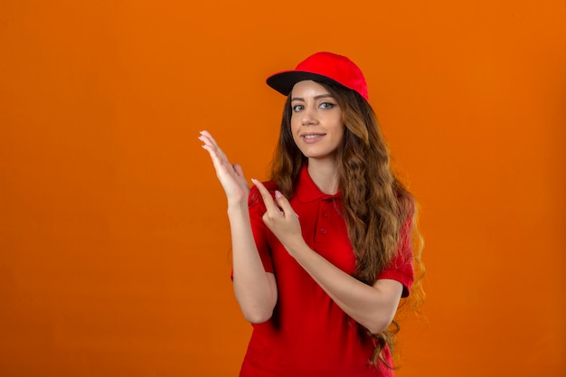 Repartidor joven vistiendo polo rojo y gorra sonriendo a la cámara mientras presenta con la mano y apunta con el dedo sobre fondo naranja aislado
