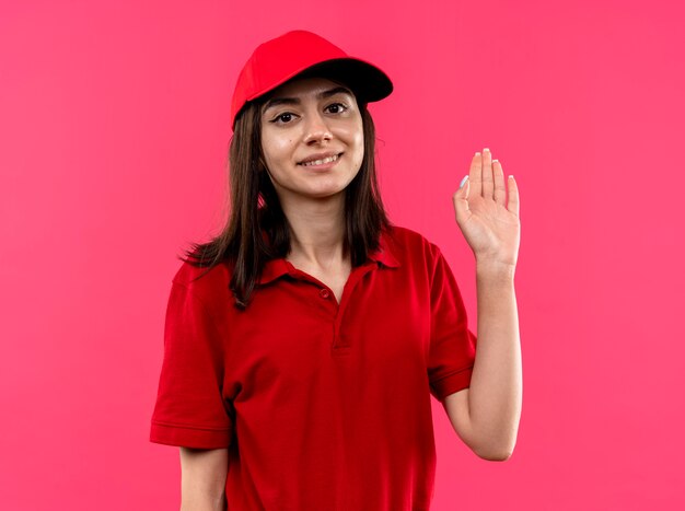 Repartidor joven vistiendo polo rojo y gorra sonriendo con cara feliz saludando con la mano de pie sobre la pared rosa