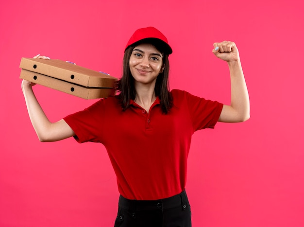 Repartidor joven vistiendo polo rojo y gorra sosteniendo cajas de pizza levantando el puño feliz y positivo sonriente amable de pie sobre fondo rosa