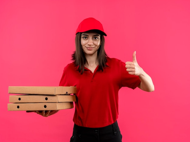 Repartidor joven vistiendo polo rojo y gorra sosteniendo cajas de pizza sonriendo mostrando los pulgares para arriba sobre la pared rosa