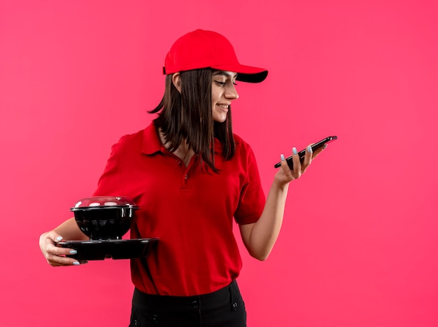 Foto gratuita repartidor joven vistiendo polo rojo y gorra sosteniendo el paquete de alimentos mirando la pantalla de su teléfono inteligente sonriendo de pie sobre la pared rosa