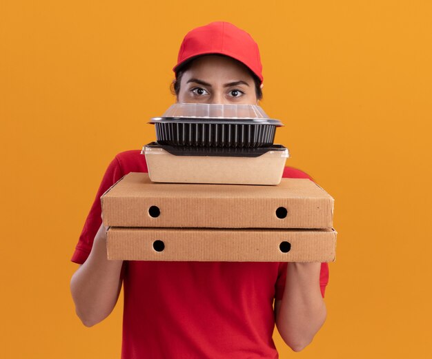 Repartidor joven vistiendo uniforme y gorra con la cara cubierta con cajas de pizza y recipientes de comida aislado en la pared naranja