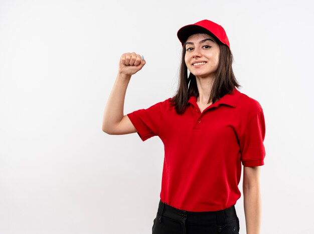 Repartidor joven vistiendo uniforme rojo y gorra apretando el puño feliz y positivo sonriendo confiado de pie sobre la pared blanca