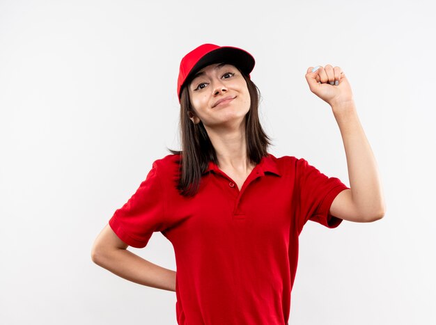 Repartidor joven vistiendo uniforme rojo y gorra apretando el puño feliz y positivo sonriendo de pie sobre la pared blanca