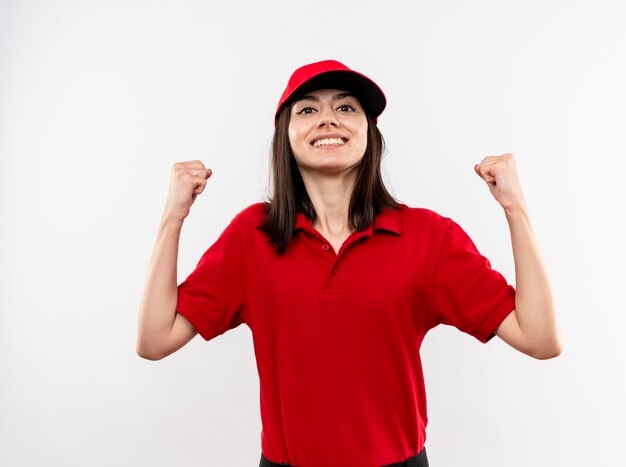 Repartidor joven vistiendo uniforme rojo y gorra mirando a la cámara apretando los puños feliz y emocionado sonriendo alegremente de pie sobre fondo blanco.