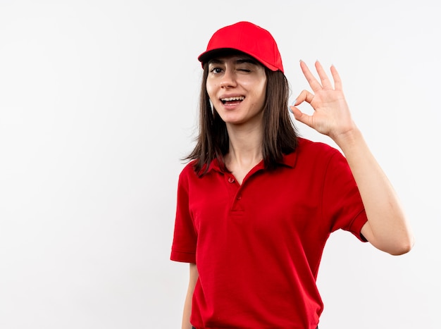 Repartidor joven vistiendo uniforme rojo y gorra mirando a la cámara sonriendo con cara feliz guiñando un ojo mostrando signo ok de pie sobre fondo blanco.