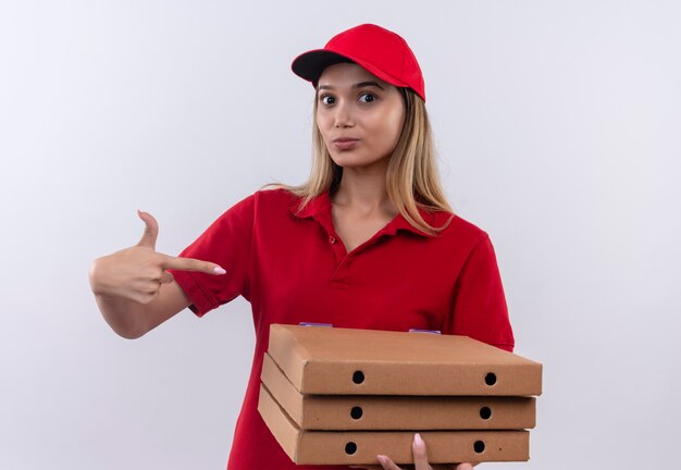 Repartidor joven vistiendo uniforme rojo y gorra sosteniendo y apunta a cajas de pizza aislado en la pared blanca