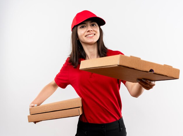 Repartidor joven vistiendo uniforme rojo y gorra sosteniendo cajas de pizza ofreciendo uno de ellos al cliente sonriendo con cara feliz de pie sobre fondo blanco.