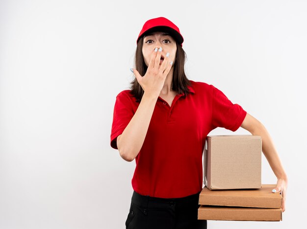 Repartidor joven vistiendo uniforme rojo y gorra sosteniendo el paquete de caja y cajas de pizza mirando a la cámara siendo sorprendido cubriendo la boca con la mano de pie sobre fondo blanco.