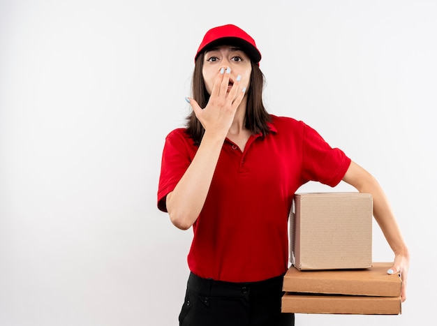 Repartidor joven vistiendo uniforme rojo y gorra sosteniendo el paquete de caja y cajas de pizza mirando a la cámara siendo sorprendido cubriendo la boca con la mano de pie sobre fondo blanco.