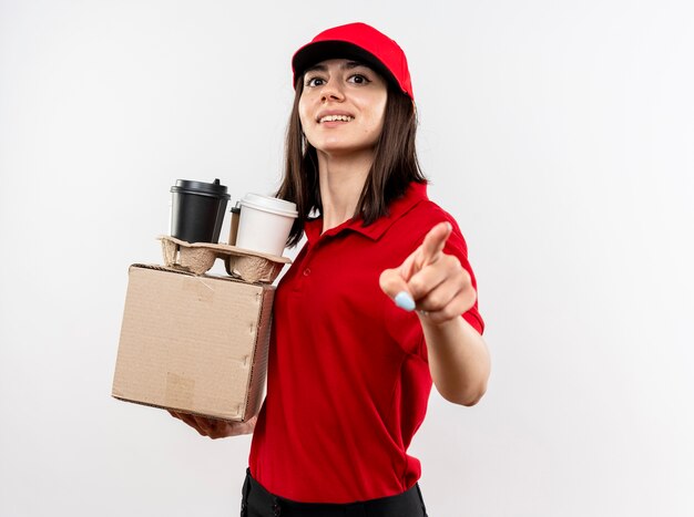 Repartidor joven vistiendo uniforme rojo y gorra sosteniendo el paquete de caja y tazas de café apuntando con el dedo índice una sonrisa con cara feliz de pie sobre una pared blanca