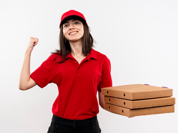 Repartidor joven vistiendo uniforme rojo y gorra sosteniendo la pila de cajas de pizza apretando el puño feliz y positivo sonriendo de pie sobre fondo blanco