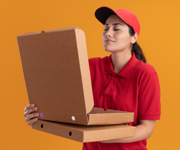 Repartidor joven vistiendo uniforme y tapa abriendo y oliendo caja de pizza aislada en pared naranja