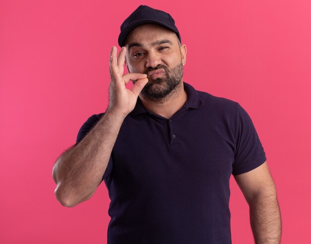 Repartidor de mediana edad complacido en uniforme y gorra que muestra un gesto delicioso aislado en la pared rosa