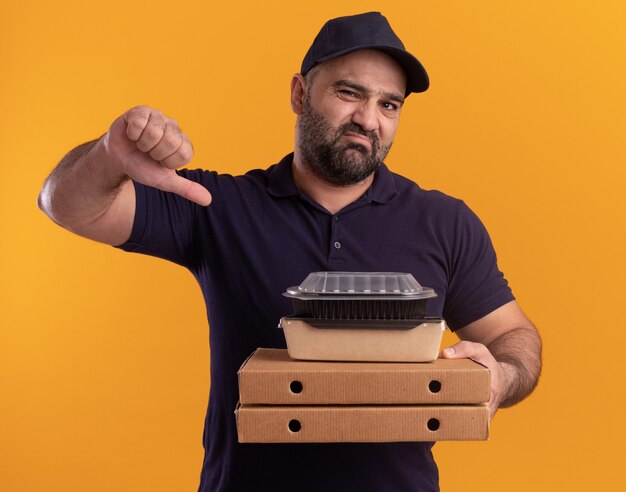 Repartidor de mediana edad disgustado en uniforme y gorra sosteniendo el recipiente de comida en cajas de pizza mostrando el pulgar hacia abajo aislado en la pared amarilla