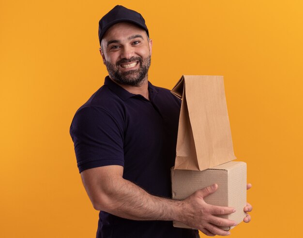 Repartidor de mediana edad sonriente en uniforme y caja de retención de tapa con paquete de comida de papel aislado en la pared amarilla