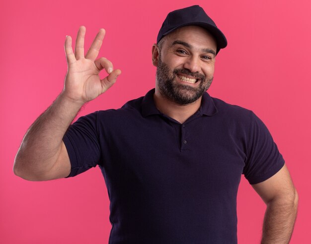 Repartidor de mediana edad sonriente en uniforme y gorra mostrando gesto bien aislado en la pared rosa