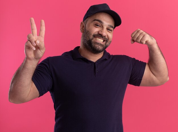 Repartidor de mediana edad sonriente en uniforme y gorra mostrando gesto de paz y haciendo un gesto fuerte aislado en la pared rosa