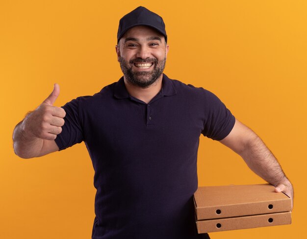 Repartidor de mediana edad sonriente en uniforme y gorra sosteniendo cajas de pizza mostrando el pulgar hacia arriba aislado en la pared amarilla