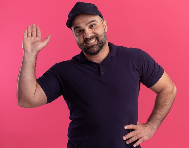 Repartidor de mediana edad sonriente en uniforme y gorra sosteniendo la mano en la cadera y mostrando gesto de saludo aislado en la pared rosa