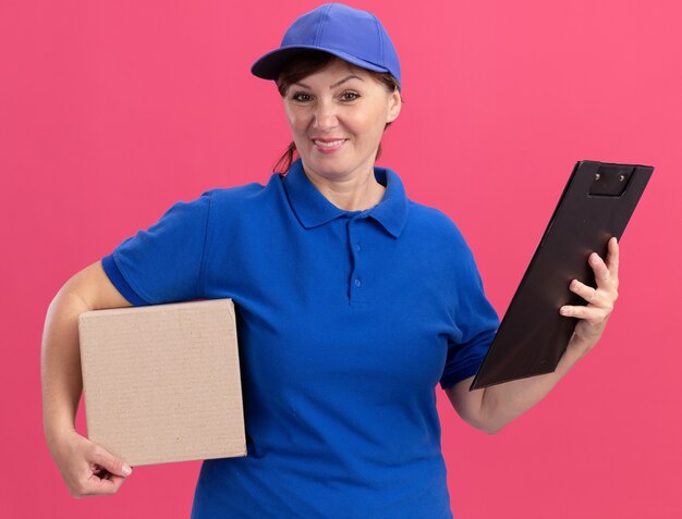 Repartidor de mediana edad en uniforme azul y gorra con caja de cartón y portapapeles mirando al frente sonriendo con cara feliz de pie sobre la pared rosa