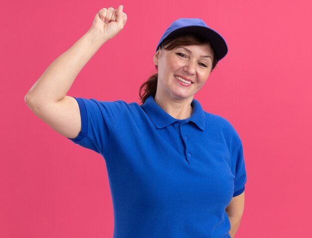 Repartidor de mediana edad en uniforme azul y gorra mirando al frente sonriendo alegremente mostrando el dedo índice de pie sobre la pared rosa