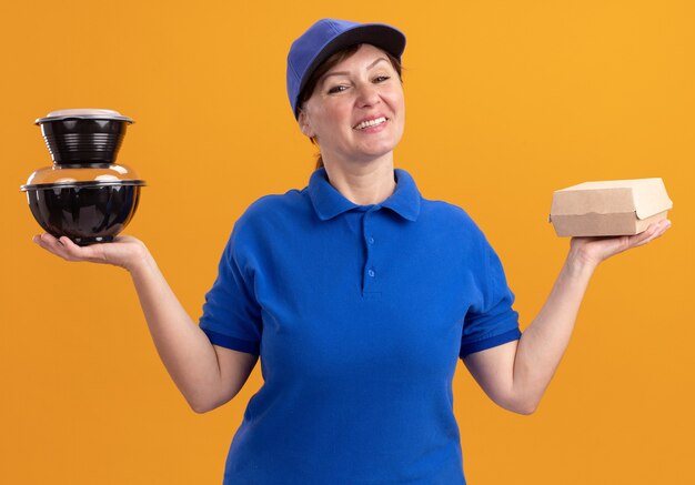 Repartidor de mediana edad en uniforme azul y gorra sosteniendo paquetes de alimentos mirando al frente sonriendo feliz y positivo de pie sobre la pared naranja