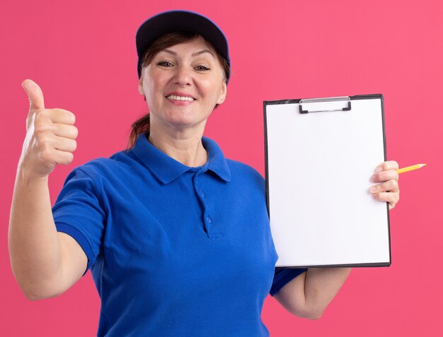 Repartidor de mediana edad en uniforme azul y gorra sosteniendo portapapeles con páginas en blanco mirando al frente sonriendo alegremente mostrando los pulgares para arriba de pie sobre la pared rosa