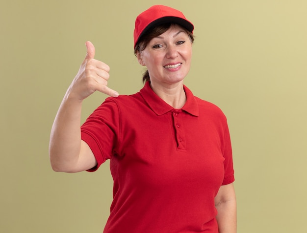 Repartidor de mediana edad en uniforme rojo y gorra mirando al frente sonriendo alegremente haciendo gesto de llamarme de pie sobre la pared verde