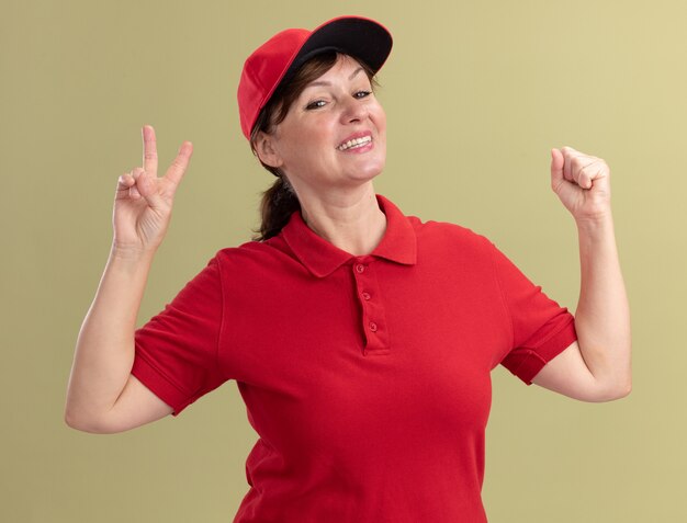 Repartidor de mediana edad con uniforme rojo y gorra mirando al frente sonriendo con cara feliz mostrando el signo v y levantando el puño de pie sobre la pared verde