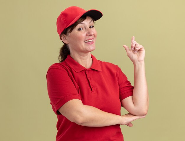 Repartidor de mediana edad con uniforme rojo y gorra mirando al frente sonriendo confiado mostrando el dedo índice con una nueva gran idea de pie sobre la pared verde