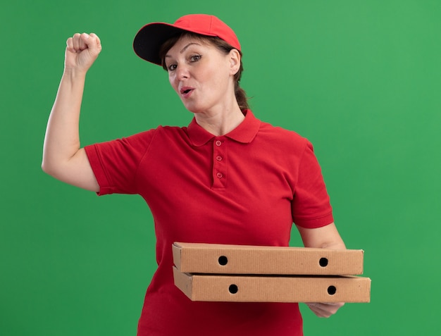 Repartidor de mediana edad en uniforme rojo y gorra sosteniendo cajas de pizza mirando al frente levantando el puño feliz y seguro de pie sobre la pared verde