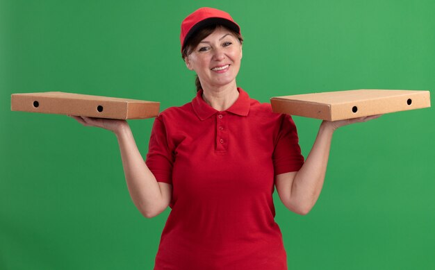 Repartidor de mediana edad en uniforme rojo y gorra sosteniendo cajas de pizza mirando al frente sonriendo con cara feliz de pie sobre la pared verde