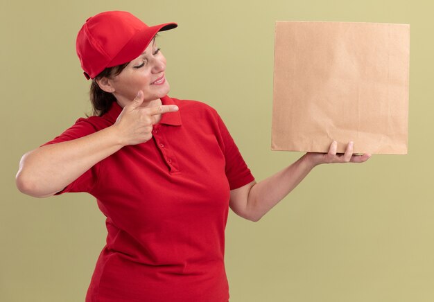 Repartidor de mediana edad en uniforme rojo y gorra sosteniendo el paquete de papel apuntando con el dedo índice sonriendo confiado de pie sobre la pared verde