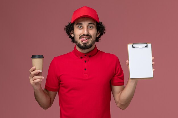 Repartidor de mensajería masculino de vista frontal en camisa roja y capa sosteniendo una taza de café marrón y un bloc de notas en la pared de color rosa claro.