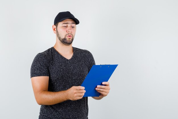 Repartidor mirando notas en el portapapeles en camiseta y gorra y mirando con cuidado