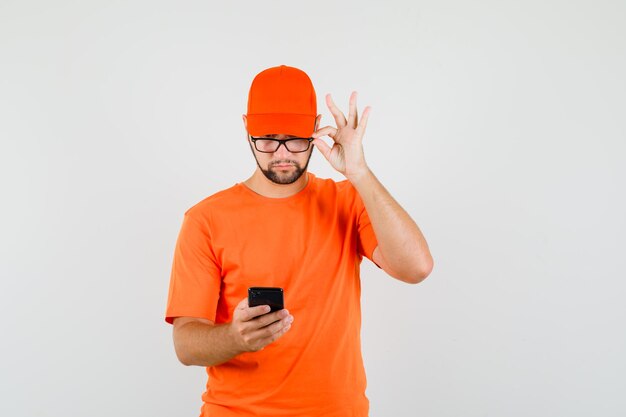 Repartidor mirando el teléfono móvil a través de gafas en camiseta naranja, gorra, vista frontal.