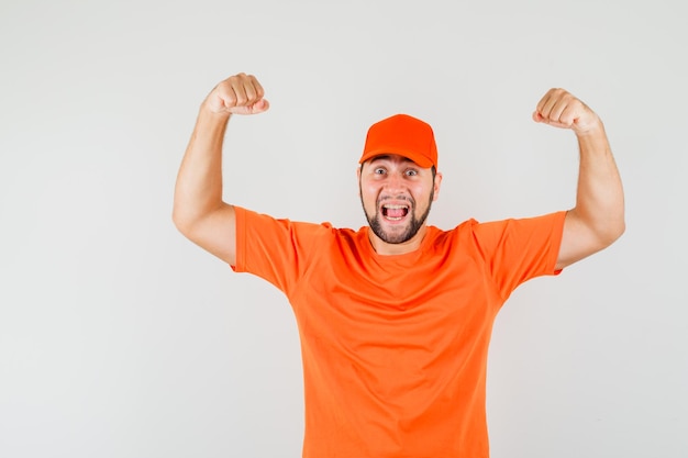 Foto gratuita repartidor mostrando gesto de éxito en camiseta naranja, gorra y mirando dichoso, vista frontal.